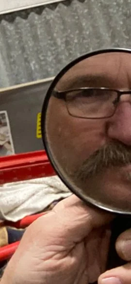 Chad caught on the other side of a magnifying glass in the shop — close-up, glasses and mustache, eyes on the work.