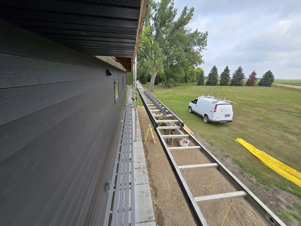 The Ranch Remodel — long view down the finished charcoal LP siding wall with scaffold plank, work van and prairie tree line beyond.